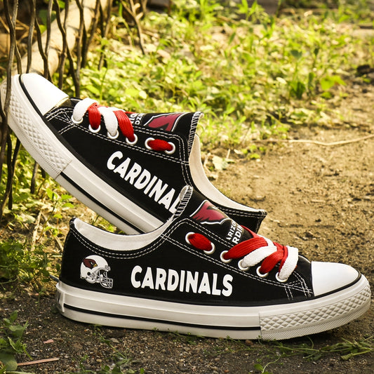 Black Arizona Cardinals canvas sneakers resting against a wire fence and grass. Highlights the white "CARDINALS" lettering and Cardinals helmet logo.
