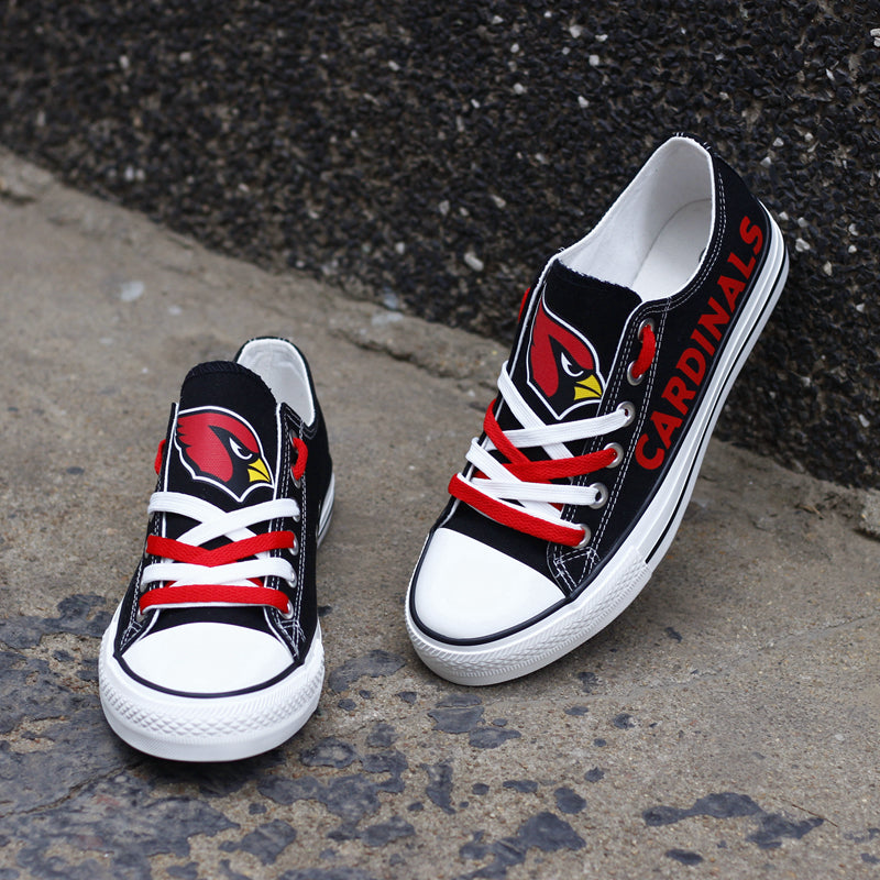One large and one small black Arizona Cardinals canvas sneaker leaning against a stone wall outdoors. Shows the red laces and the iconic cardinal head logo.