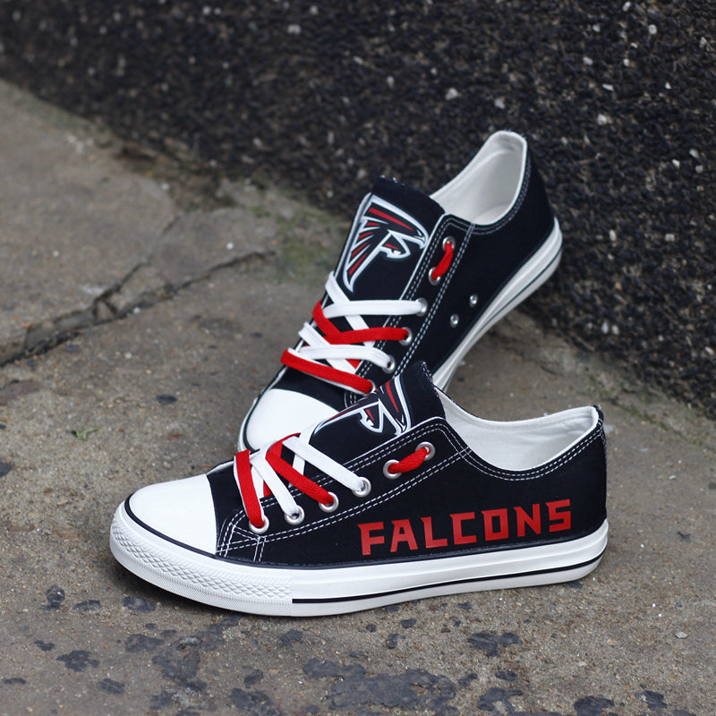 Pair of black Atlanta Falcons canvas sneakers resting against a dark textured wall outdoors. Focuses on the red 'FALCONS' text and the stylish look for urban fan wear.