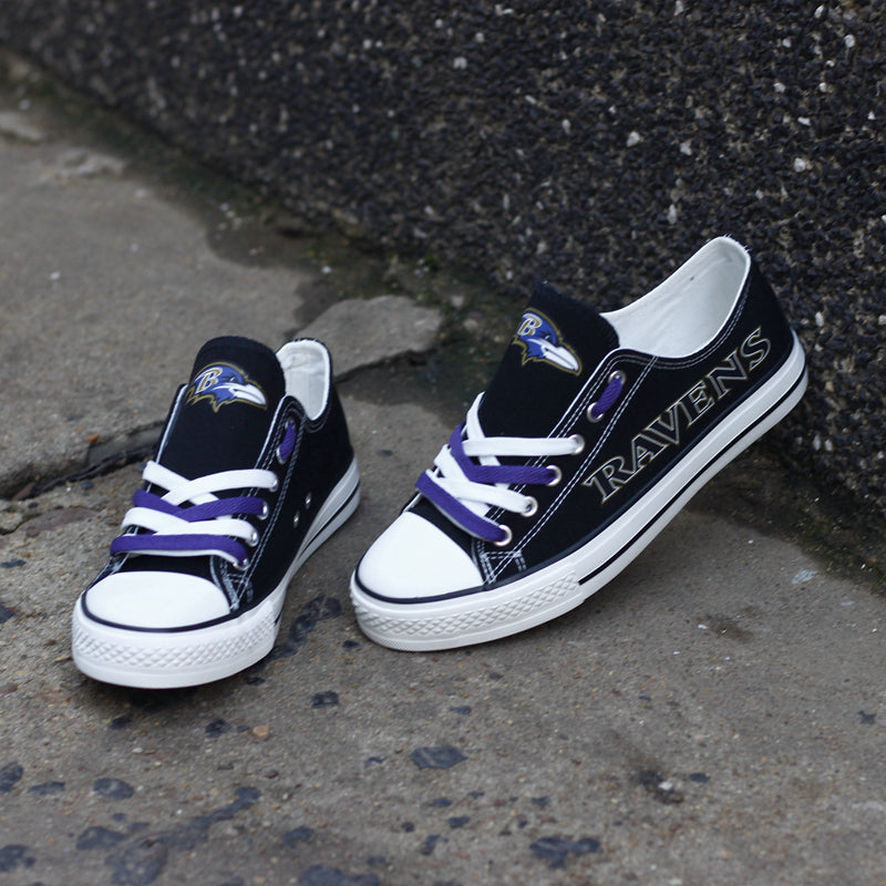 A pair of black Baltimore Ravens canvas shoes with white soles and purple/white laces, resting against a textured concrete wall. Casual NFL fan footwear.
