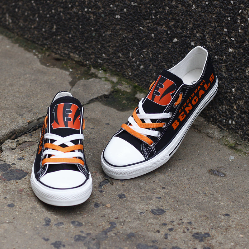 A pair of black Cincinnati Bengals canvas shoes on a concrete surface, angled upward. Focuses on the orange laces and the team logo on the tongue.