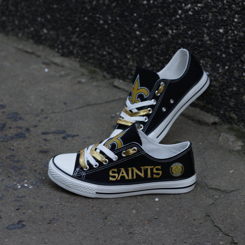 Black New Orleans Saints canvas sneakers with gold text and fleur-de-lis, photographed outdoors on a rough concrete background.