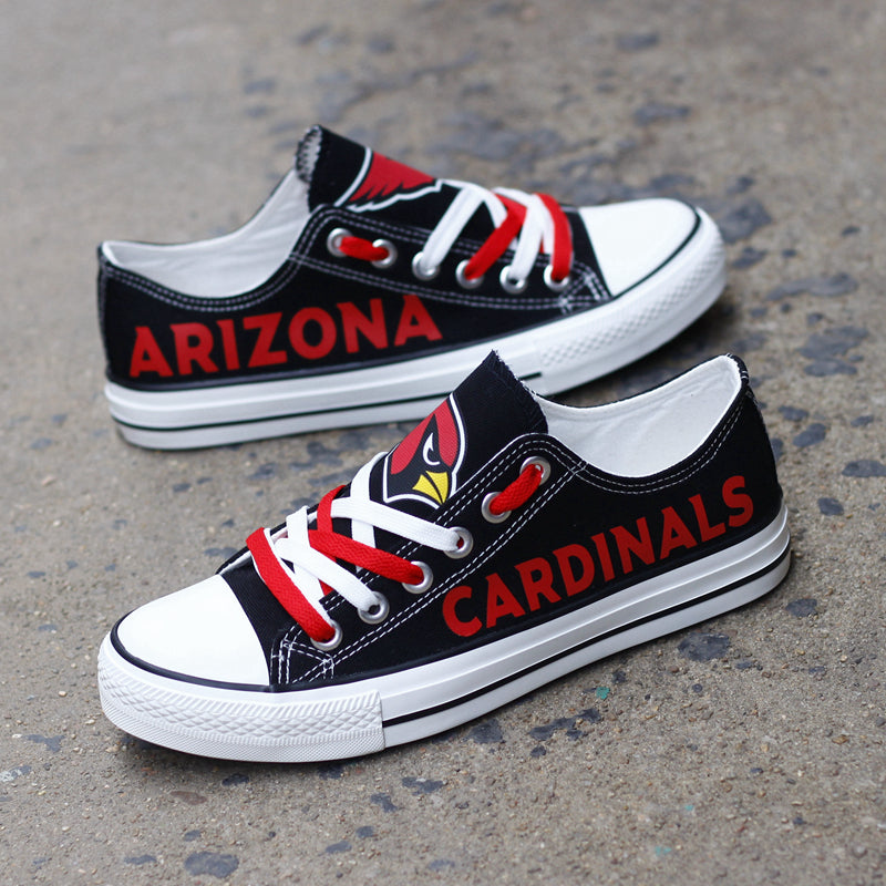 Pair of black Arizona Cardinals canvas low-top shoes on a gray concrete surface. Features the vibrant red 'CARDINALS' lettering and the classic team emblem.