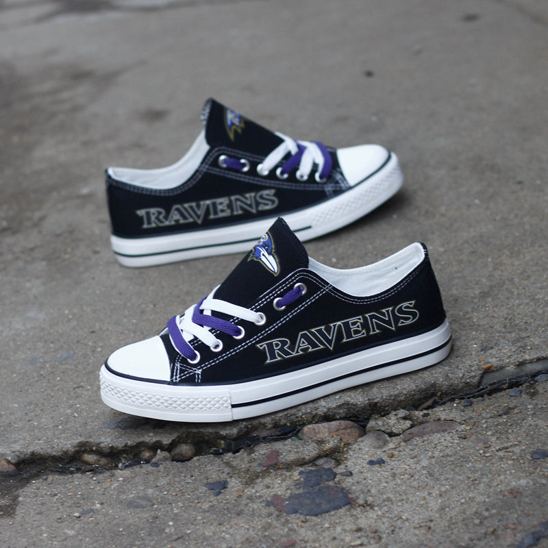 Two black Baltimore Ravens canvas sneakers displayed outdoors on a cracked concrete surface. Shoes have purple accents and are ready for everyday wear or tailgating.