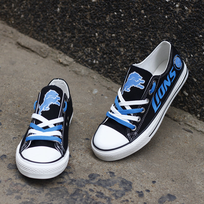 One large and one small black Detroit Lions canvas sneaker leaning against a stone wall outdoors. Shows the blue laces and the iconic lion head logo.