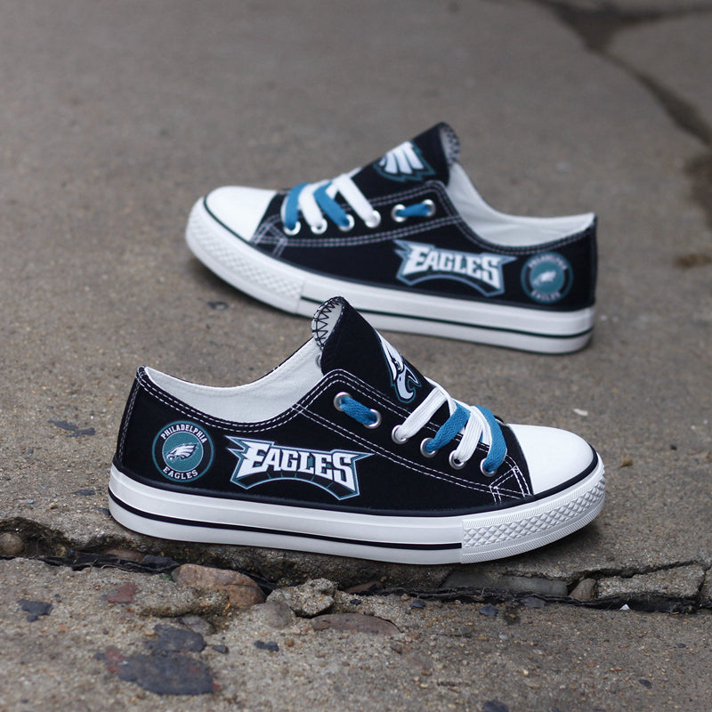 Two black Philadelphia Eagles canvas low-top shoes sitting on a cracked concrete sidewalk. Features the white and teal 'EAGLES' lettering clearly.