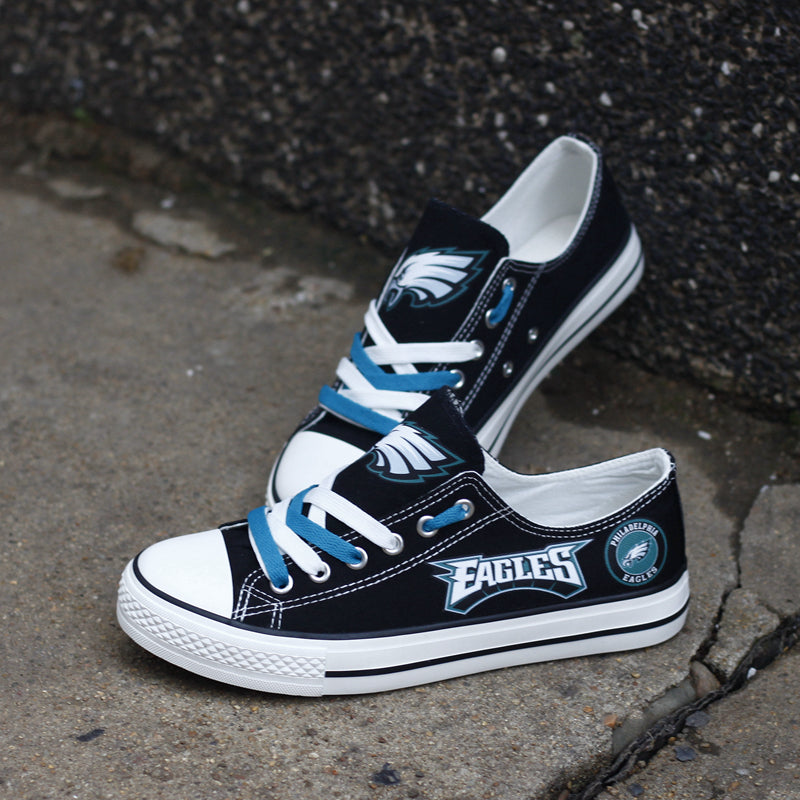 Black Philadelphia Eagles canvas low-tops leaning against a dark textured wall outdoors. Highlights the bold 'EAGLES' logo and teal accent colors.
