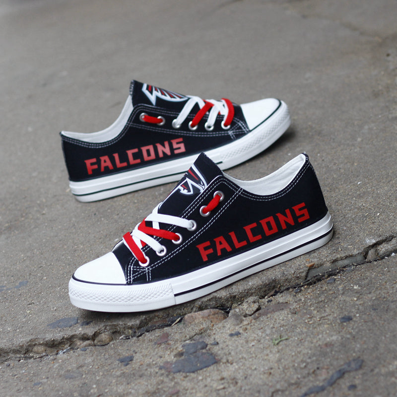 Pair of black Atlanta Falcons canvas low-top shoes on a gray concrete surface. Features the vibrant red 'FALCONS' lettering and the classic team emblem.