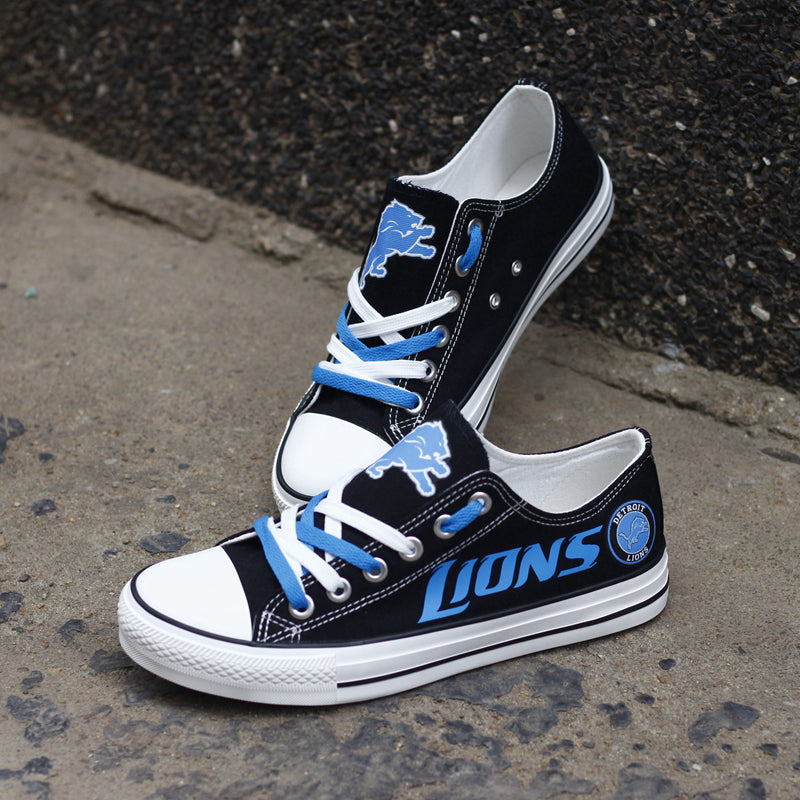 Black Detroit Lions canvas sneakers resting against a dark textured wall outdoors. Highlights the blue 'LIONS' lettering and the lion head logo for an urban fan vibe.