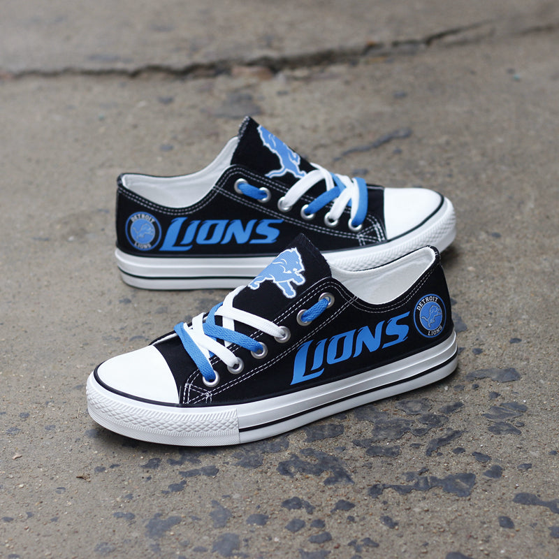 Pair of black Detroit Lions canvas low-top shoes on a gray concrete surface. Features the vibrant blue 'LIONS' lettering and the classic team emblem.