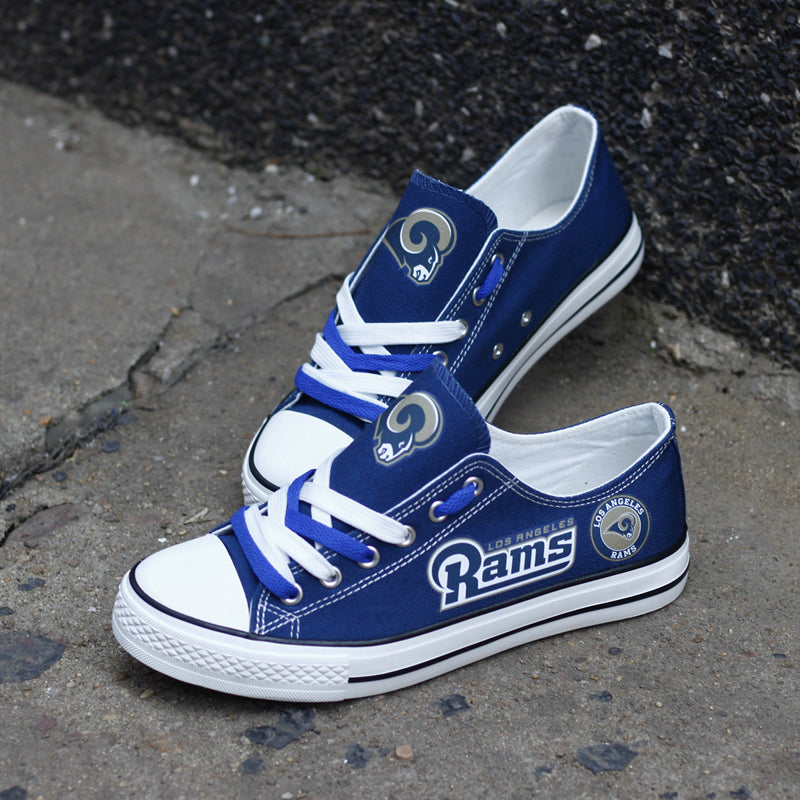 Pair of royal blue Los Angeles Rams canvas low-top shoes on a gray concrete surface. Shows the vibrant gold 'RAMS' lettering and the team logo on the tongue.