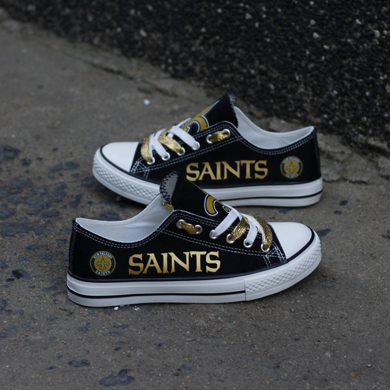 Two New Orleans Saints black and gold canvas sneakers resting on a dark concrete street surface, highlighting the "SAINTS" lettering.