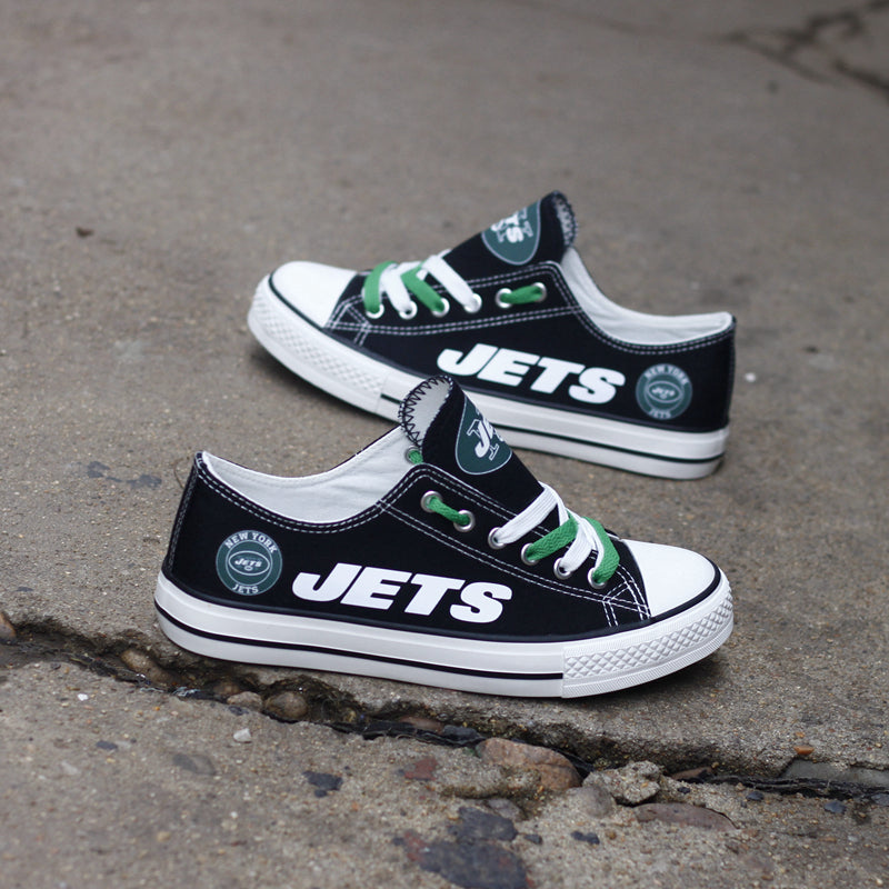 Pair of black New York Jets canvas low-top shoes on a gray concrete surface. Shows the vibrant white 'JETS' lettering and the round team logo.