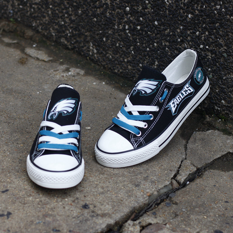 A small and large black Philadelphia Eagles canvas shoe on an outdoor concrete surface. Shows the team logo and the teal laces.