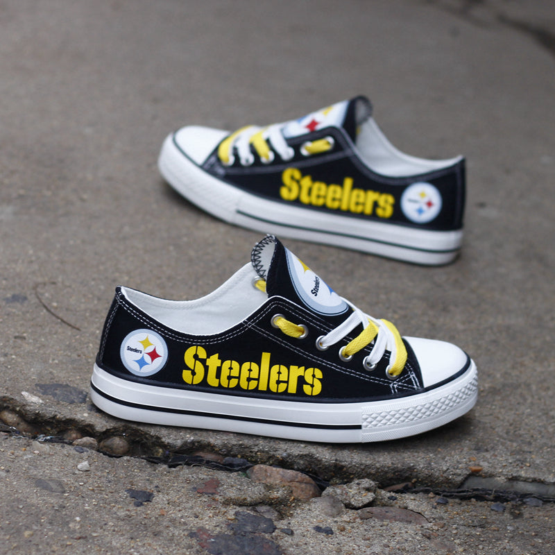 Pair of black Pittsburgh Steelers canvas low-top shoes on a gray concrete surface. Shows the vibrant yellow 'Steelers' lettering and the three-diamond logo.