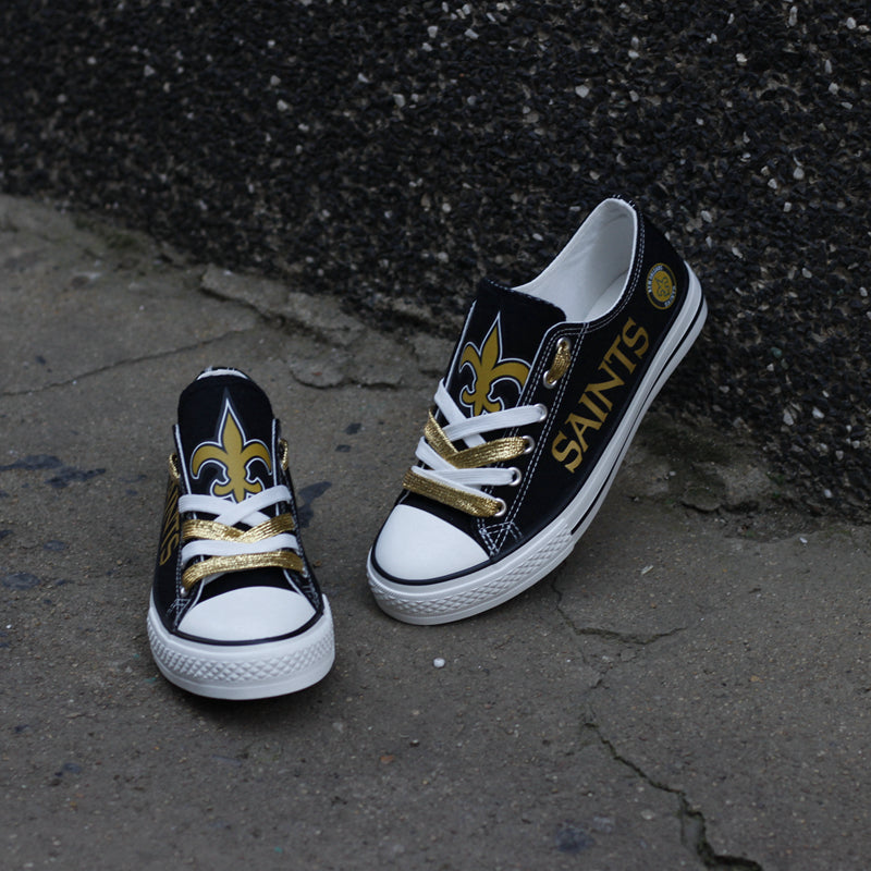 One New Orleans Saints canvas shoe standing against a dark wall and one on the ground, showcasing the gold fleur-de-lis logo and "SAINTS" text.