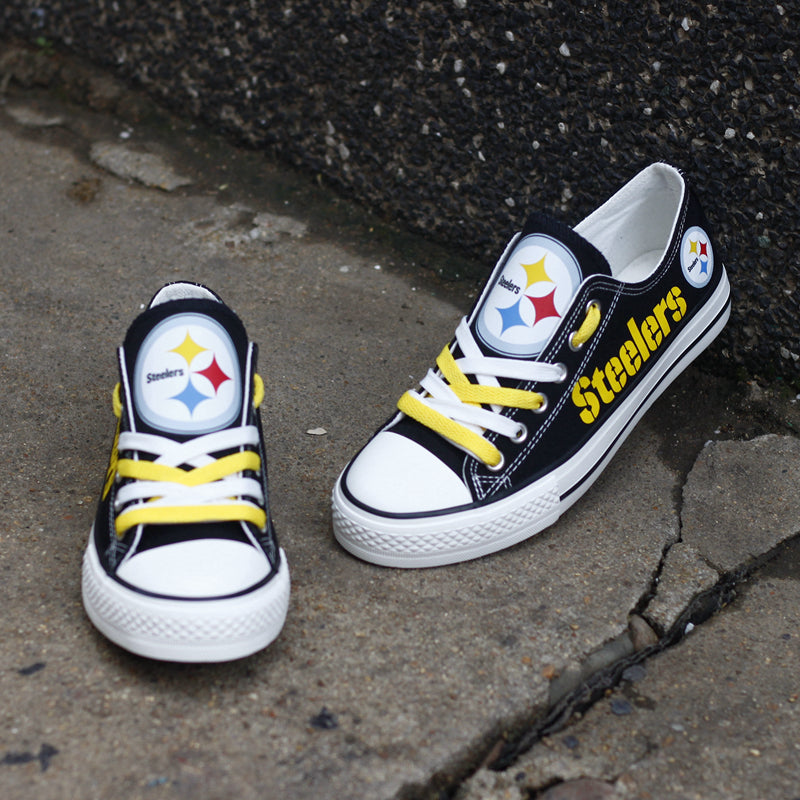 One large and one small black Pittsburgh Steelers canvas sneaker resting on concrete against a dark wall. Highlights the yellow laces and the iconic Steeler logo on the tongue.