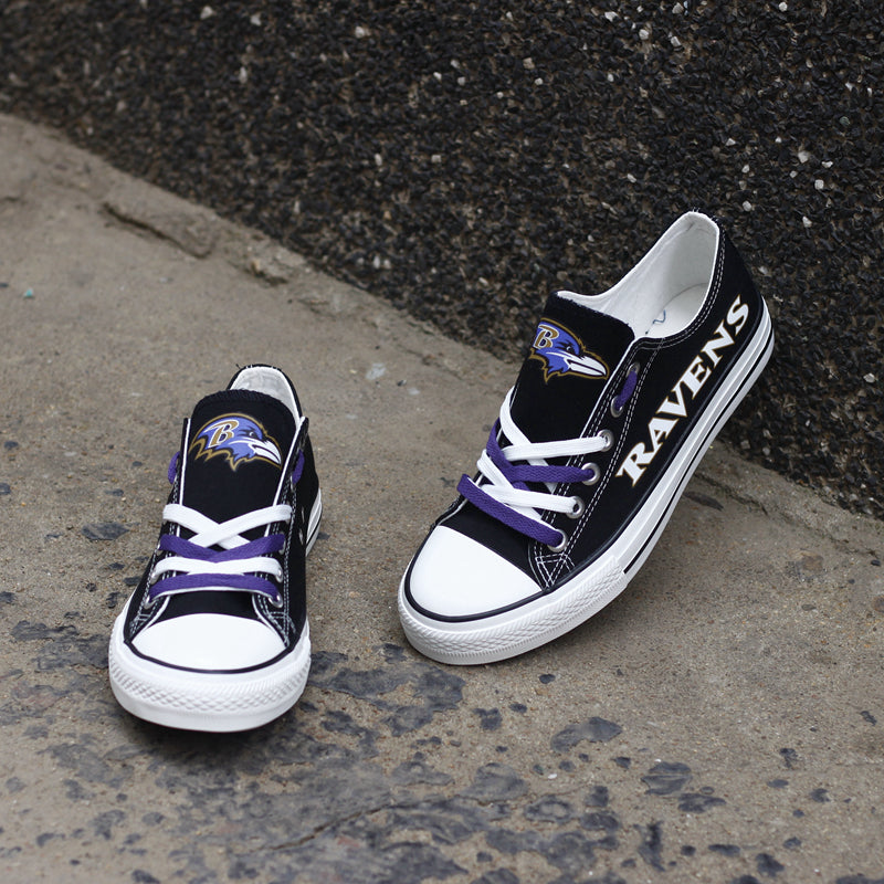 Two sizes (adult and youth/mini) of the black Baltimore Ravens canvas shoes on a concrete surface. Features the team logo and purple laces for all family members.