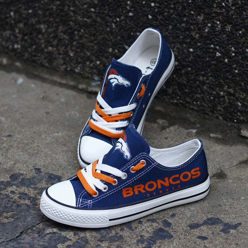 Navy blue Denver Broncos canvas sneakers resting against a dark textured wall outdoors. Highlights the orange 'BRONCOS DENVER' lettering and orange laces.
