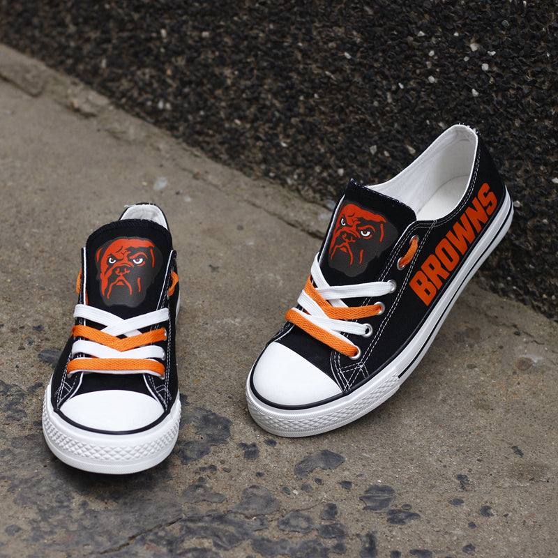 Pair of black Cleveland Browns canvas low-top shoes on a gray concrete surface. Features the vibrant orange 'BROWNS' lettering and the classic team emblem.