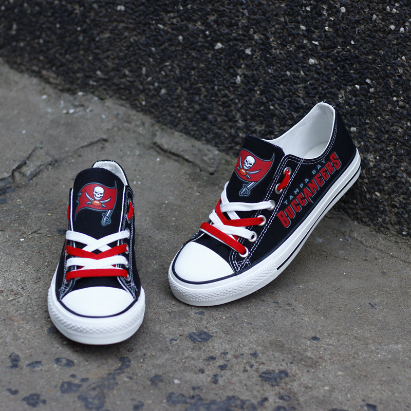 Black Tampa Bay Buccaneers canvas sneakers resting against a dark textured wall outdoors. Highlights the red 'BUCCANEERS' lettering and the pirate flag logo for an urban fan vibe.