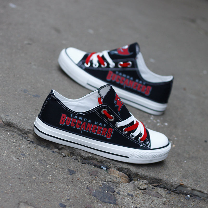 Pair of black Tampa Bay Buccaneers canvas low-top shoes on a gray concrete surface. Features the vibrant red 'BUCCANEERS' lettering and the classic team emblem.