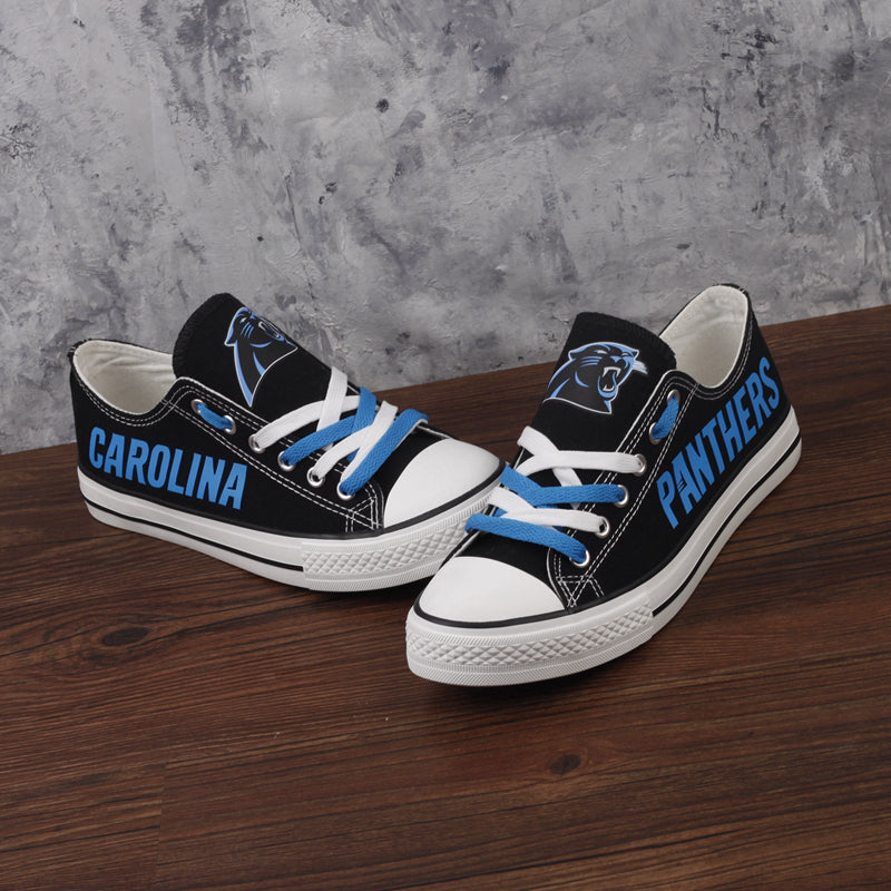 Pair of black Carolina Panthers canvas sneakers on a wooden surface. Highlights the blue 'CAROLINA' text, the team logo, and light blue laces.