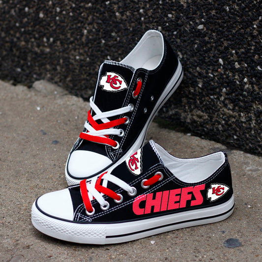 One large and one small black Kansas City Chiefs canvas sneaker leaning against a dark textured wall outdoors. Shows the red laces and the iconic team logo.