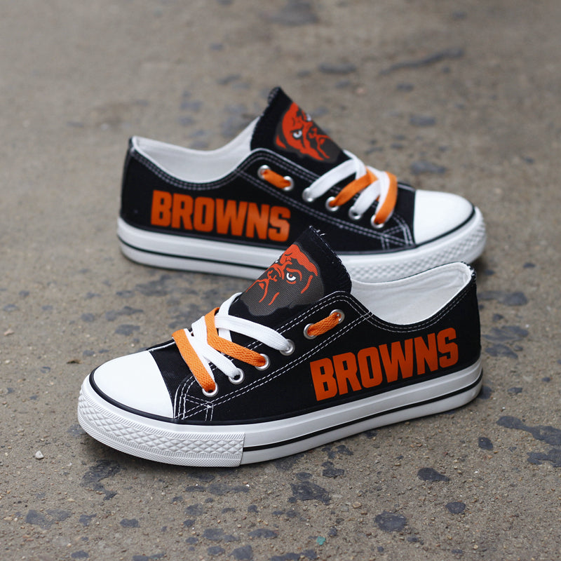 One large and one small black Cleveland Browns canvas sneaker leaning against a stone wall outdoors. Shows the orange laces and the iconic team helmet logo.