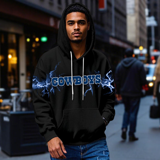 Male model wearing a black Dallas Cowboys pullover hoodie with bright blue lightning graphics, standing in a city street. The Cowboys apparel is shown being worn casually.