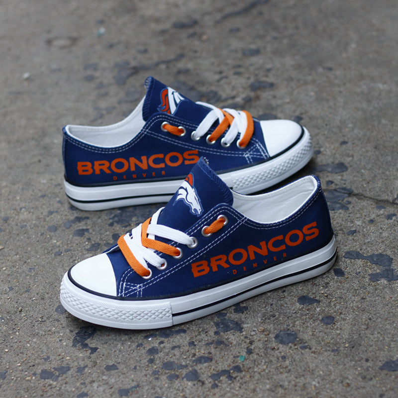 Pair of navy blue Denver Broncos canvas low-top shoes on a gray concrete surface. Features the horse logo, orange laces, and the 'BRONCOS' text.