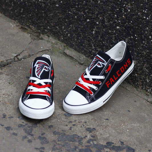 One large and one small black Atlanta Falcons canvas low-top shoe leaning against a dark textured wall outdoors. Highlights the red 'FALCONS' lettering and the team logo.