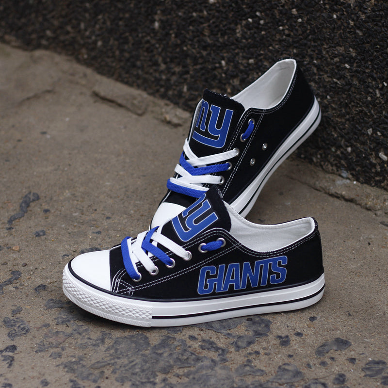 Two pairs of black New York Giants canvas low-top shoes leaning against a dark wall. Shows the blue and white laces and the iconic team logo.