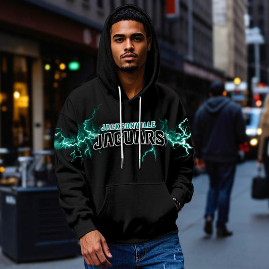 Male model wearing a black Jacksonville Jaguars pullover hoodie with bright teal lightning graphics, standing in a city street. The Jaguars apparel is shown being worn casually.