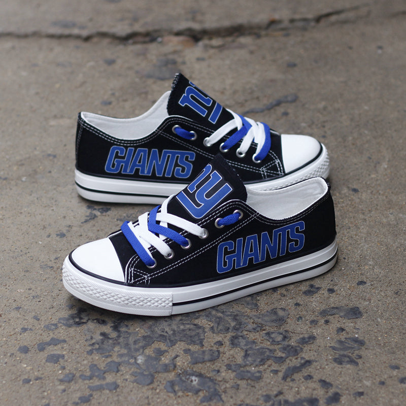 Pair of black New York Giants canvas low-top shoes on a gray concrete surface. Features the vibrant blue 'GIANTS' lettering and the classic team emblem.