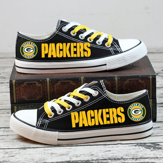 Stacked pair of black Green Bay Packers low-top canvas sneakers with yellow PACKERS text and team logo, photographed on a stack of books.