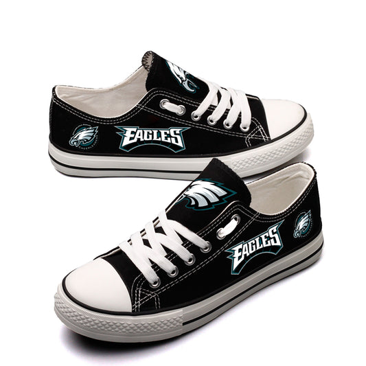 Pair of black Philadelphia Eagles canvas low-top shoes on a gray concrete surface. Shows the vibrant white 'EAGLES' lettering and the classic team logo.