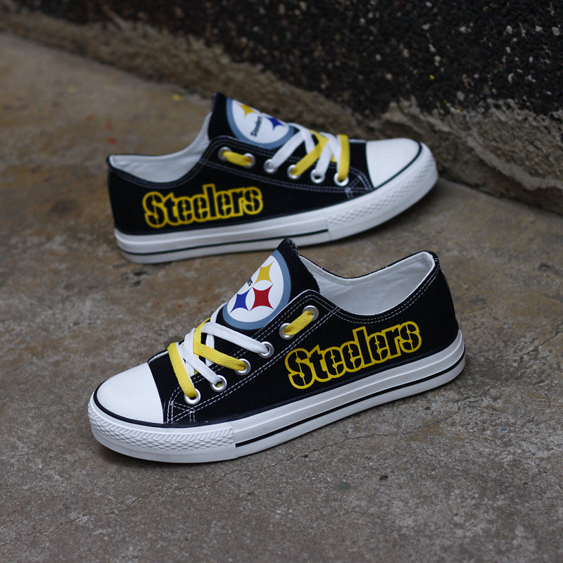Pair of black Pittsburgh Steelers canvas low-top shoes on a gray concrete surface. Features the team logo, yellow laces, and the yellow 'Steelers' text.