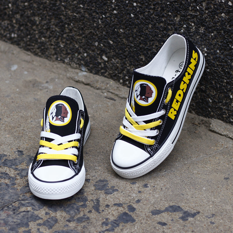 Pair of black Washington Redskins canvas low-top shoes on a gray concrete surface. Features the vibrant yellow 'REDSKINS' lettering and the classic team emblem.