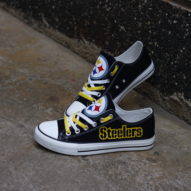 Black Pittsburgh Steelers canvas sneakers resting against a dark textured wall outdoors. Highlights the yellow 'Steelers' lettering and the yellow laces.