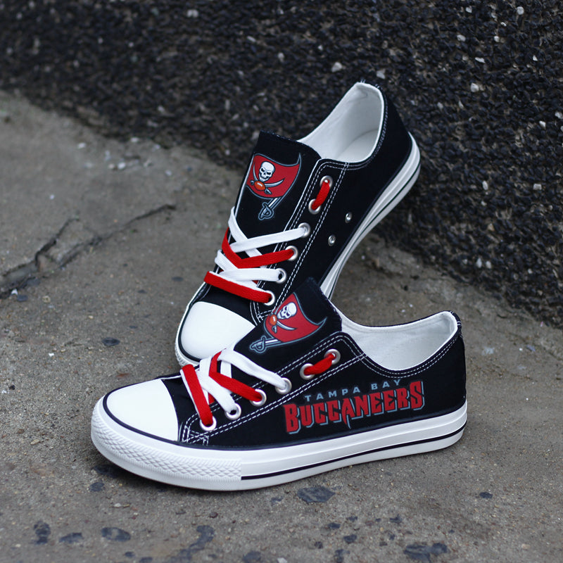 One large and one small black Tampa Bay Buccaneers canvas sneaker leaning against a dark textured wall outdoors. Shows the red laces and the iconic team logo.