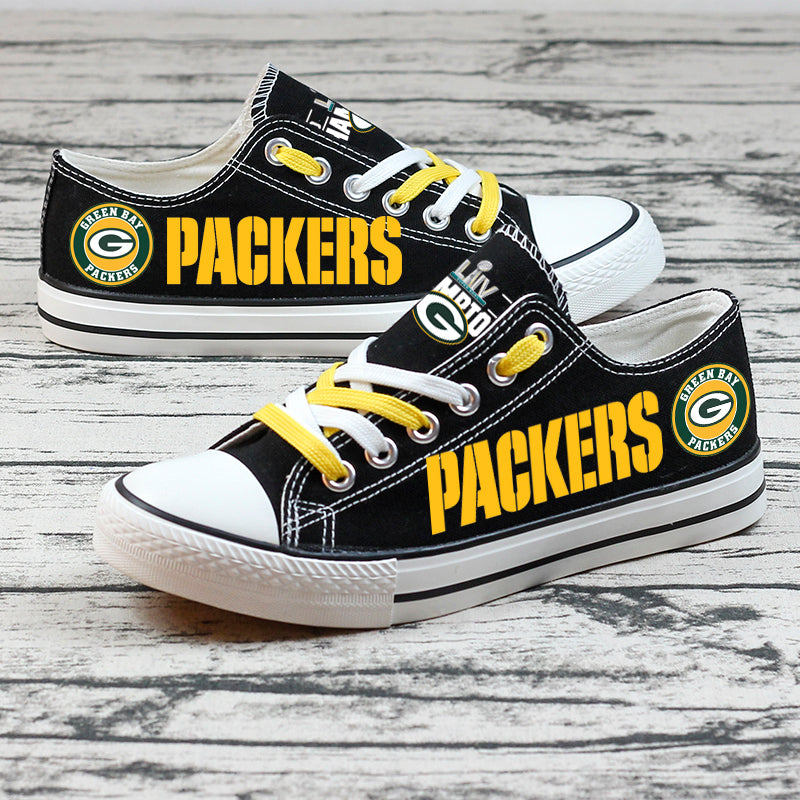 Close-up of black Green Bay Packers canvas shoes with yellow laces, team logo on the tongue, and yellow PACKERS text, set on a wooden surface.